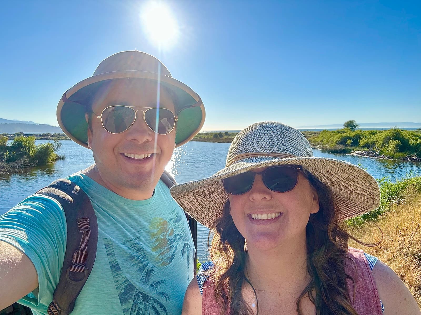 Andrew Christison and his wife, both in sun hats, on a bluff above a coastal lagoon with the Strait of Juan de Fuca in the distance.